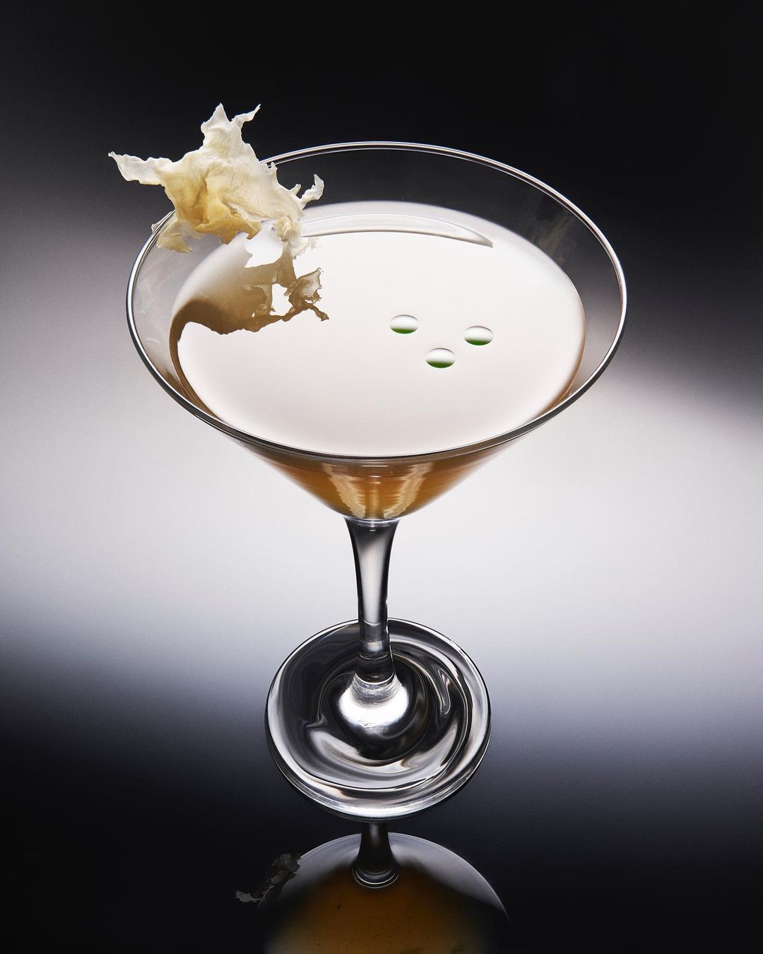 Cocktail in a martini glass garnished with an edible flower and green drops, set against a gradient black background with reflection.