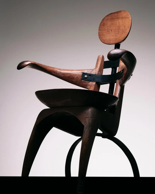 Close-up of a wooden sculptural chair with metal supports featuring a rounded back, curved armrests, and a built-in bowl seat.
