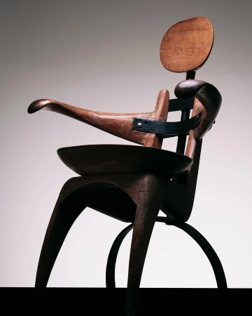 Close-up of a wooden sculptural chair with metal supports featuring a rounded back, curved armrests, and a built-in bowl seat.