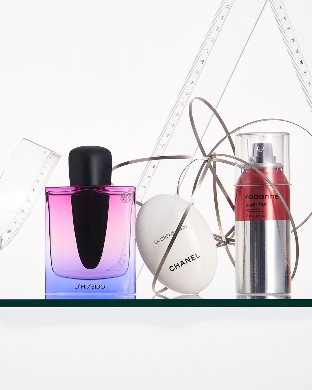 Three cosmetic product bottles: Shiseido fragrance, Chanel face cream, and Rabanne serum, arranged on a glass surface with ribbon and drafting tools.