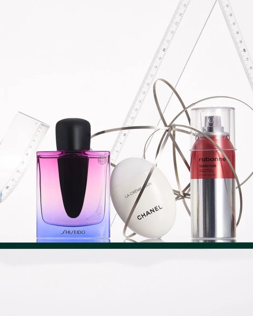Three cosmetic product bottles: Shiseido fragrance, Chanel face cream, and Rabanne serum, arranged on a glass surface with ribbon and drafting tools.