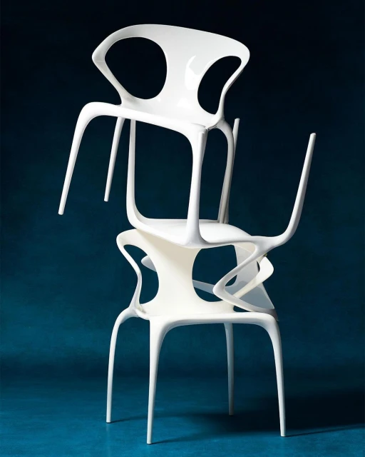 Stack of three modern, white, sculptural chairs in a studio setting against a dark blue background. Design is minimalist and artistic.
