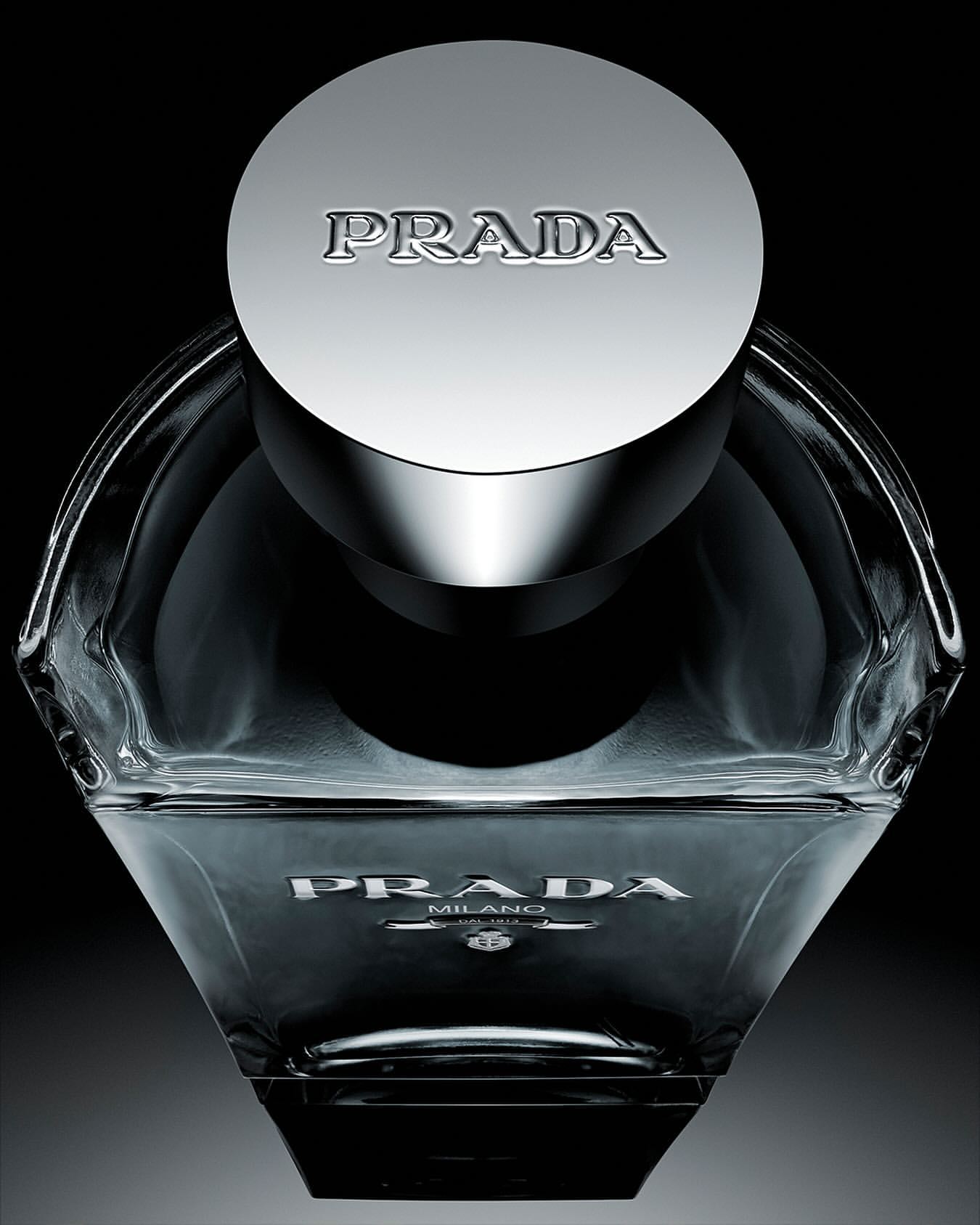 Close-up studio shot of a clear glass Prada fragrance bottle with a metallic cap against a dark background, emphasizing its design and branding.