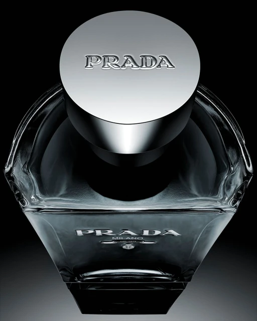 Close-up studio shot of a clear glass Prada fragrance bottle with a metallic cap against a dark background, emphasizing its design and branding.