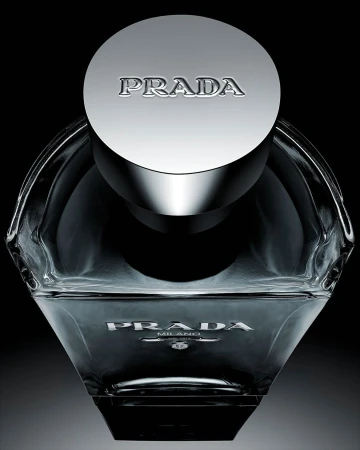 Close-up studio shot of a clear glass Prada fragrance bottle with a metallic cap against a dark background, emphasizing its design and branding.