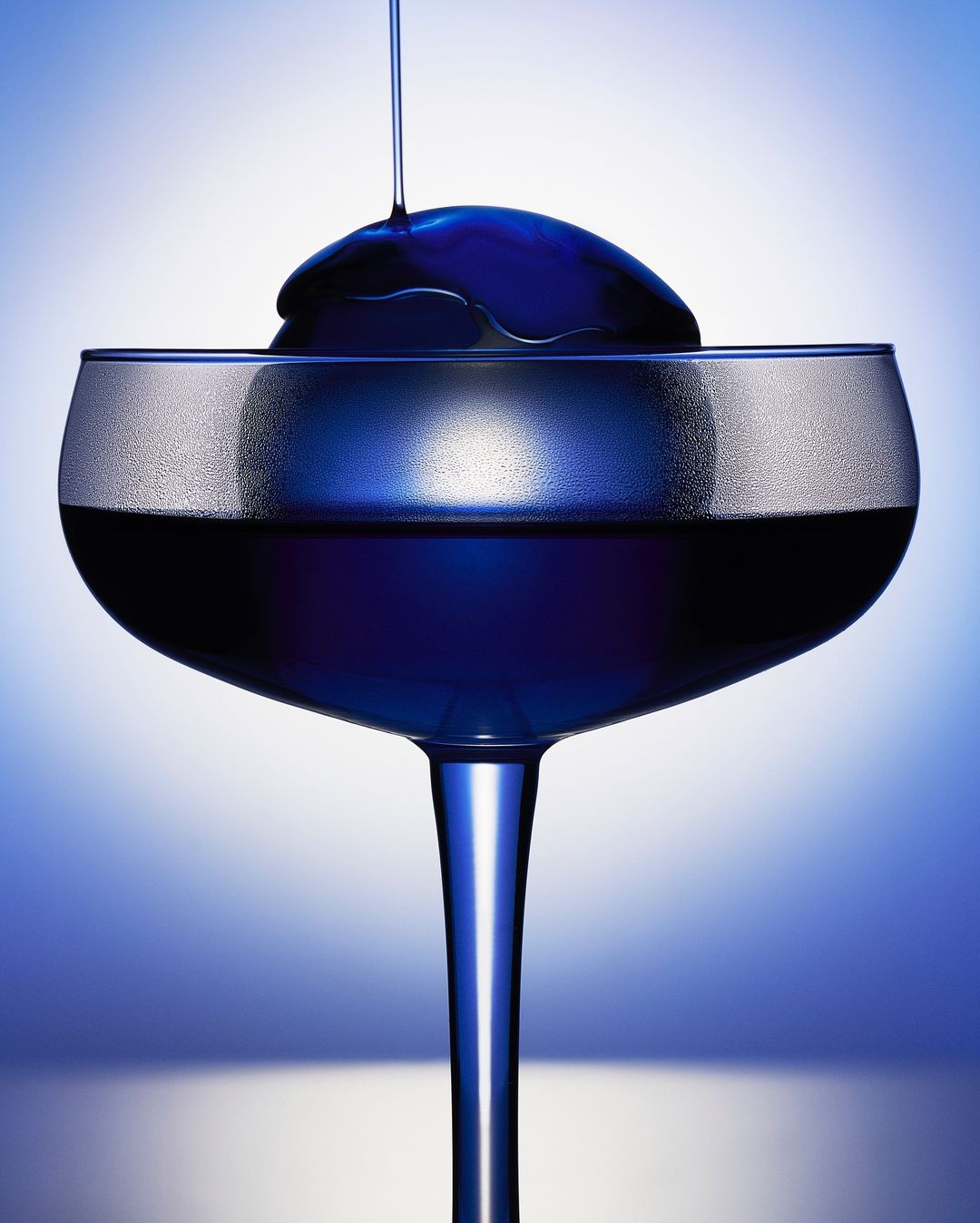 Close-up of a stemmed glass filled with blue liquid, topped with a dollop of liquid, against a graduated blue and white background.