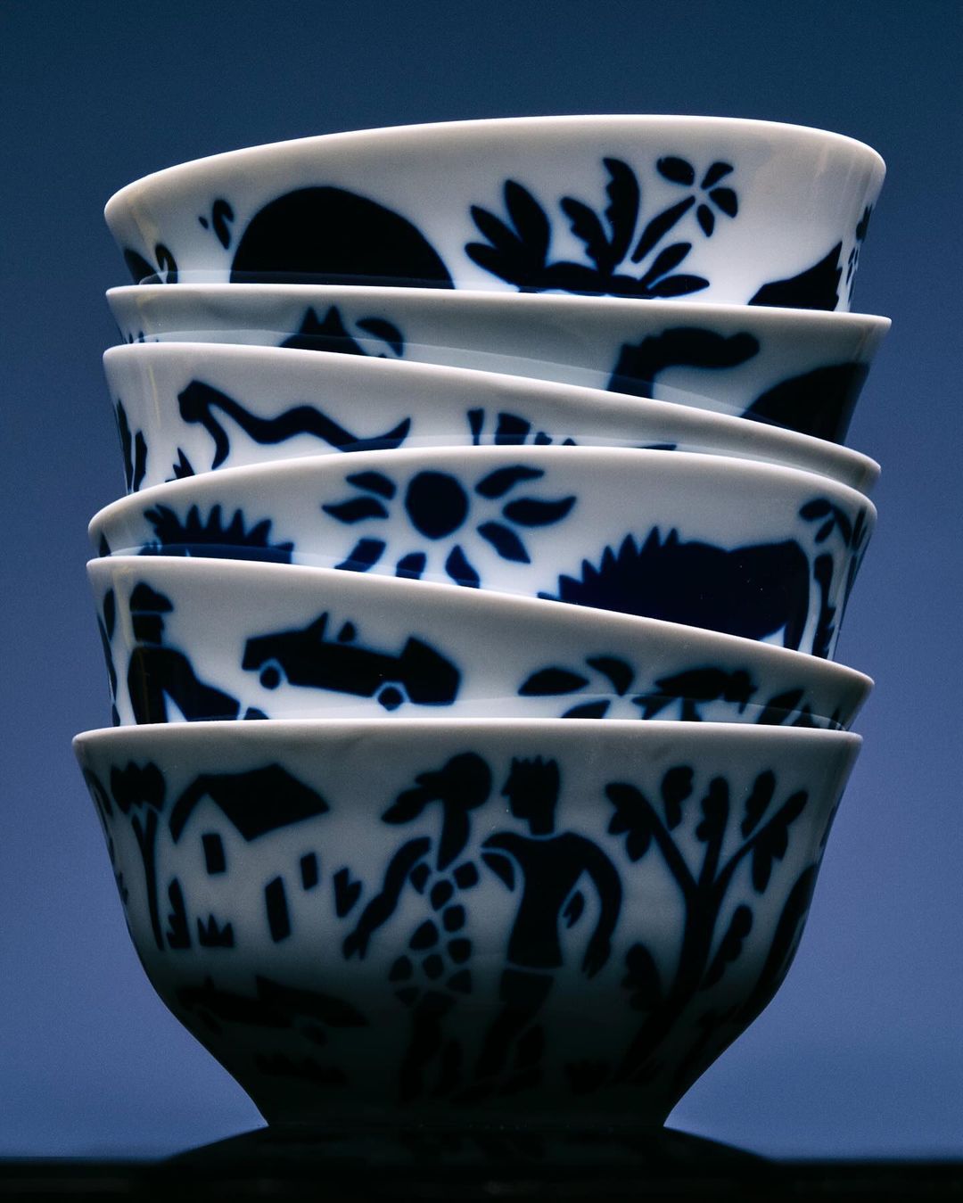 Stack of six white porcelain bowls with dark blue decorative patterns of silhouetted people, houses, animals, and nature scenes.