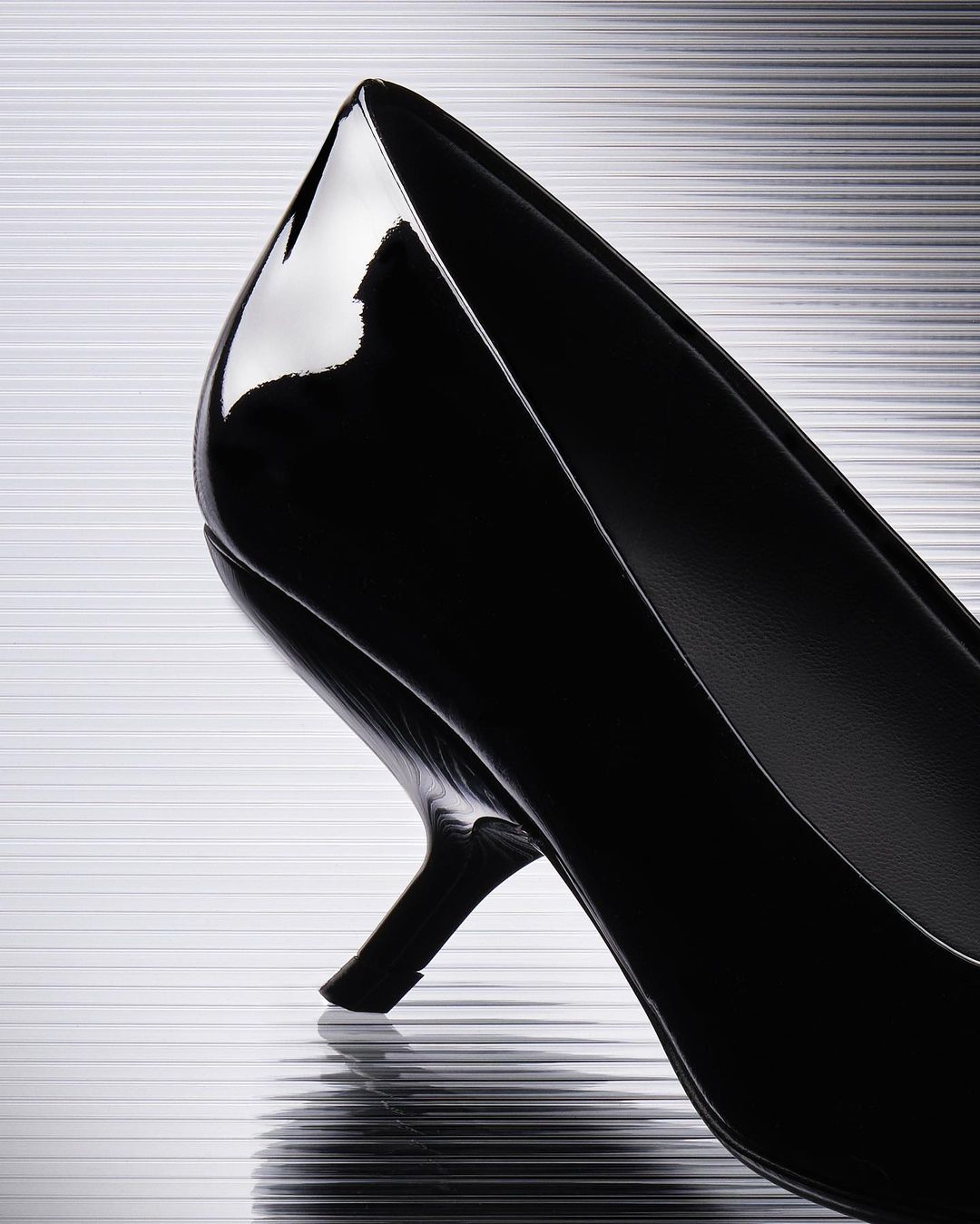 Elegant black patent leather high heel shoe on a silver horizontally striped background, showcasing fashion footwear and stylish design.