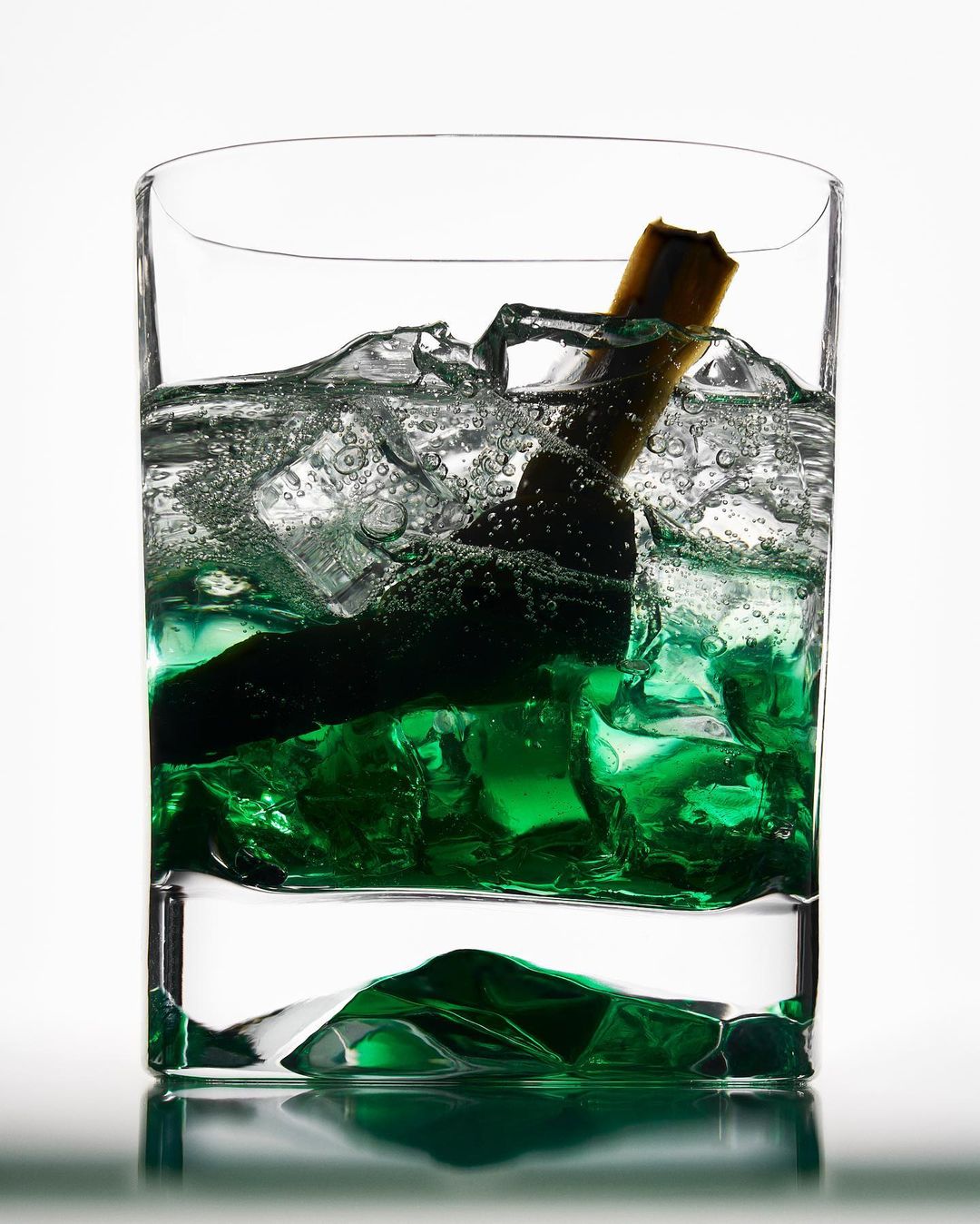 Green cocktail in a clear glass with ice cubes and a cinnamon stick garnish, set against a bright white background.