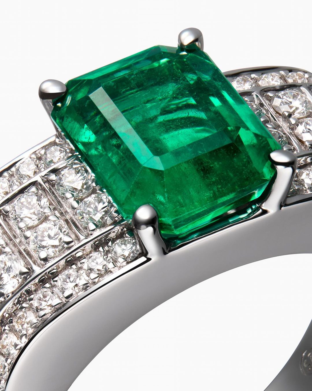 Close-up of a silver ring featuring a large, emerald-cut green emerald stone, adorned with small diamonds, on a white backdrop.