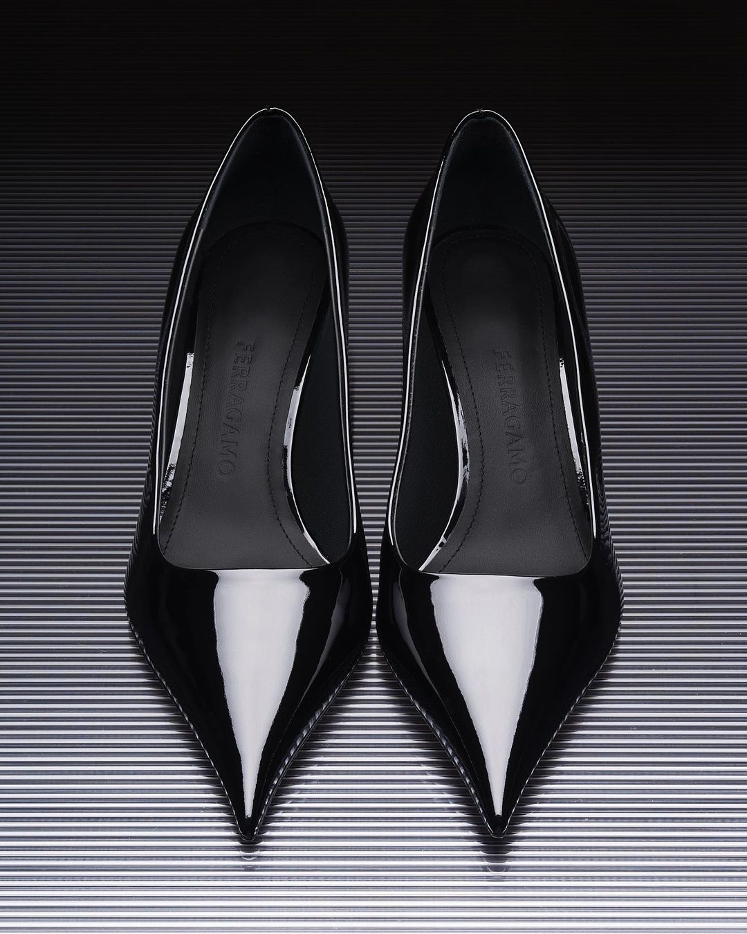 Studio product shot featuring a pair of black patent leather pointed-toe stiletto heels against a striped silver background.