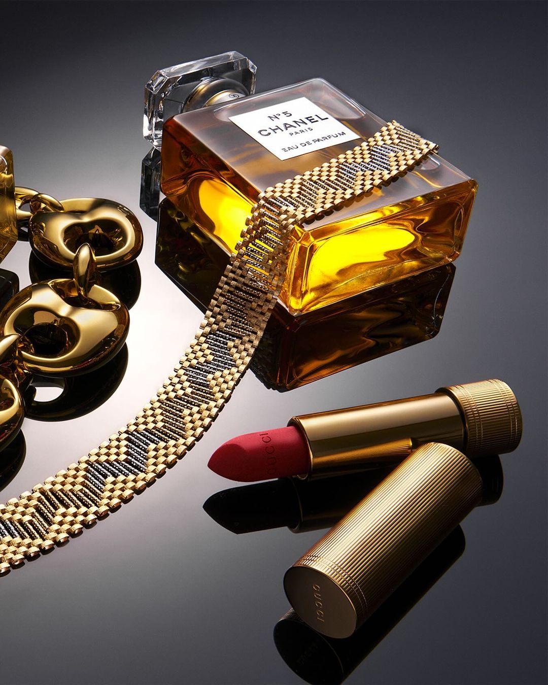 Still life shot of a Chanel perfume bottle, Gucci lipstick, and gold chain on a glossy black surface, product photography, studio lighting.