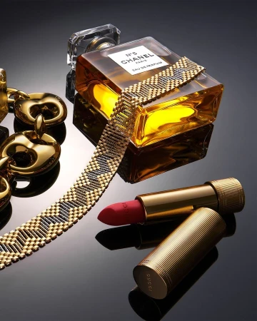 Still life shot of a Chanel perfume bottle, Gucci lipstick, and gold chain on a glossy black surface, product photography, studio lighting.