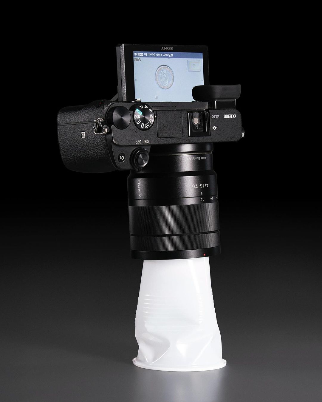 Digital camera with a large lens stands upright, using a white plastic cup as a makeshift lens hood, set against a dark backdrop.