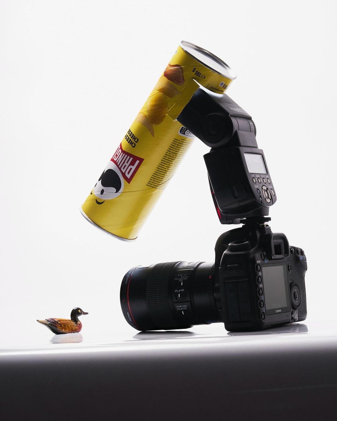 Still life studio photography featuring a digital SLR camera with flash attachment, a can of Pringles, and a toy duck on a white surface.