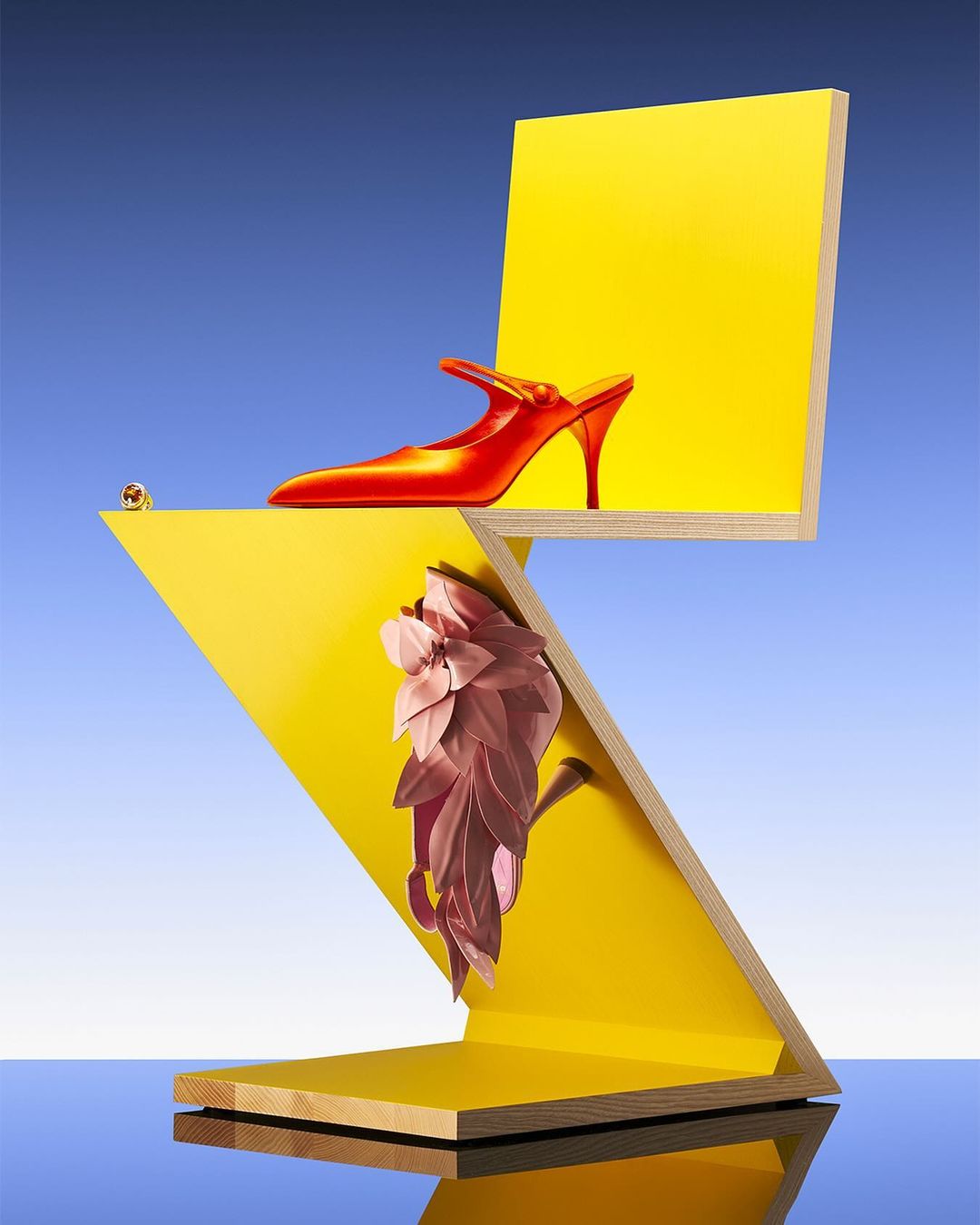 Red heeled shoe atop a yellow geometric structure against a blue sky, showcasing modern fashion and abstract design elements.