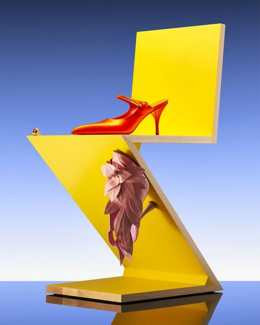 Red heeled shoe atop a yellow geometric structure against a blue sky, showcasing modern fashion and abstract design elements.