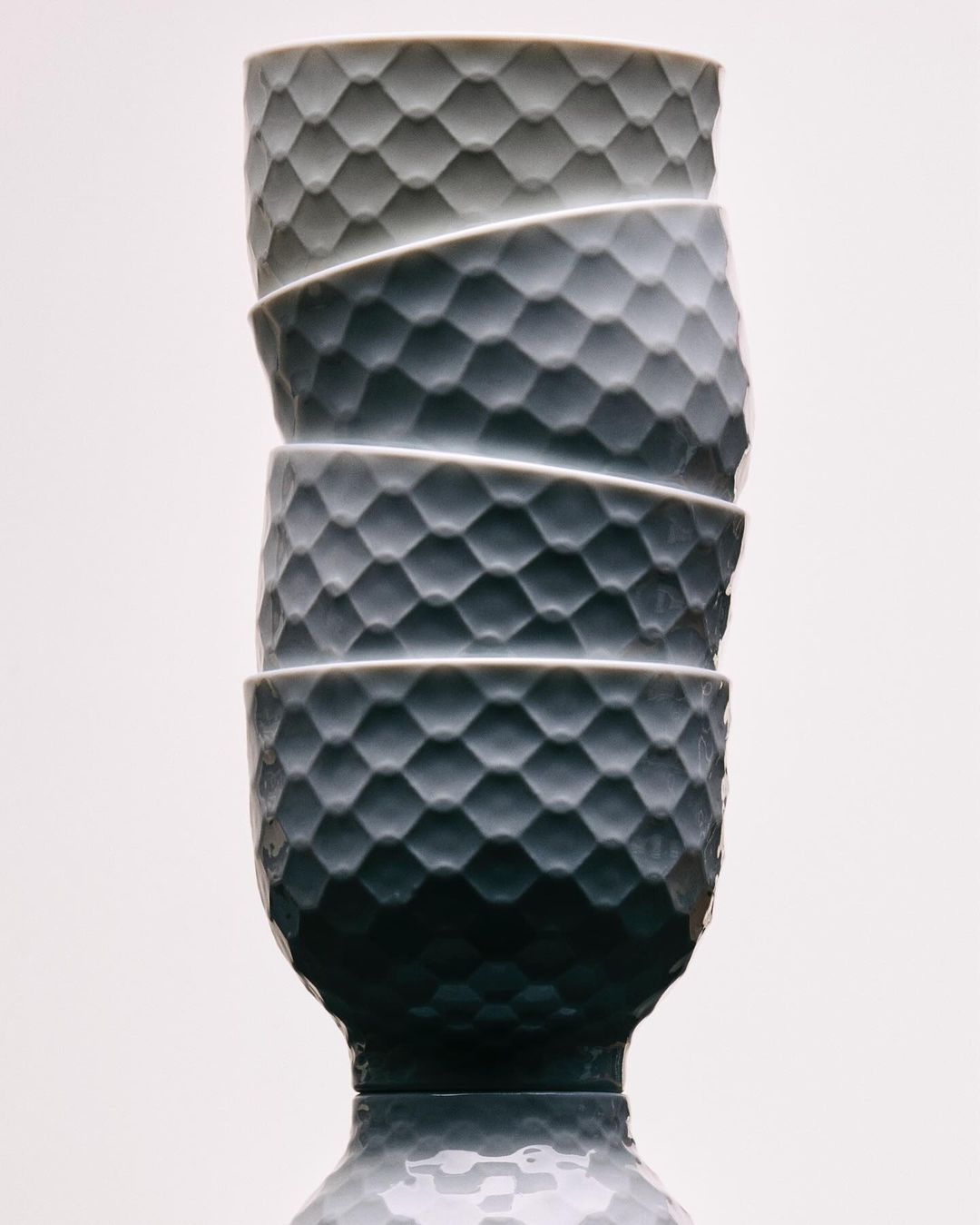 Stack of four gray ceramic bowls with a textured, faceted surface on a reflective surface against a light background in a studio setting.