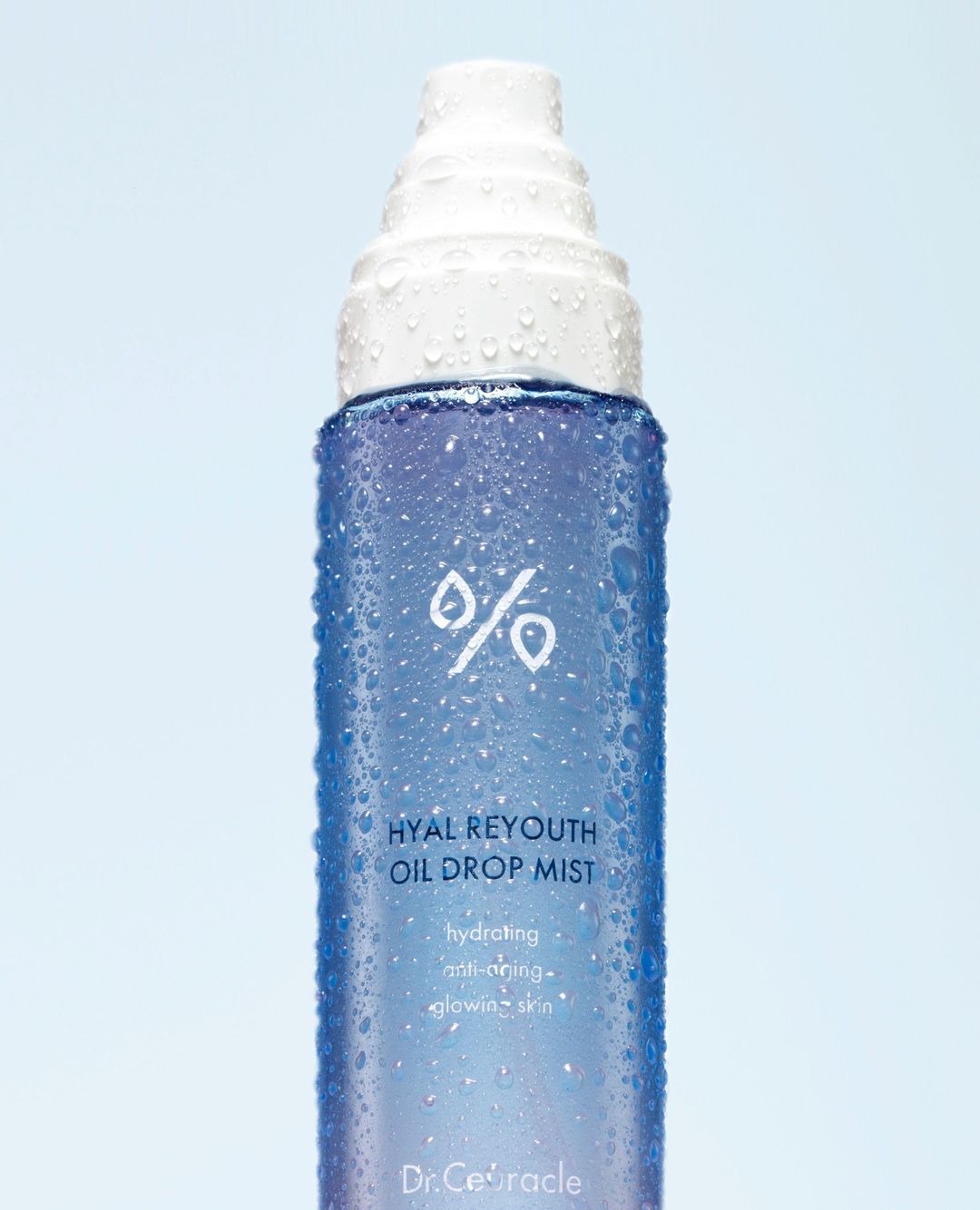 Close-up of a blue and white bottle of Hyal Reyouth Oil Drop Mist skincare product with water droplets against a light blue background.