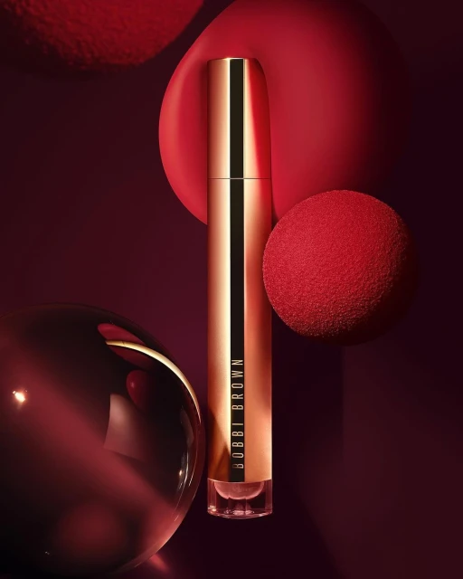 Vertical product shot of Bobbi Brown cosmetic with gold packaging, set against a backdrop of red and burgundy spherical elements.