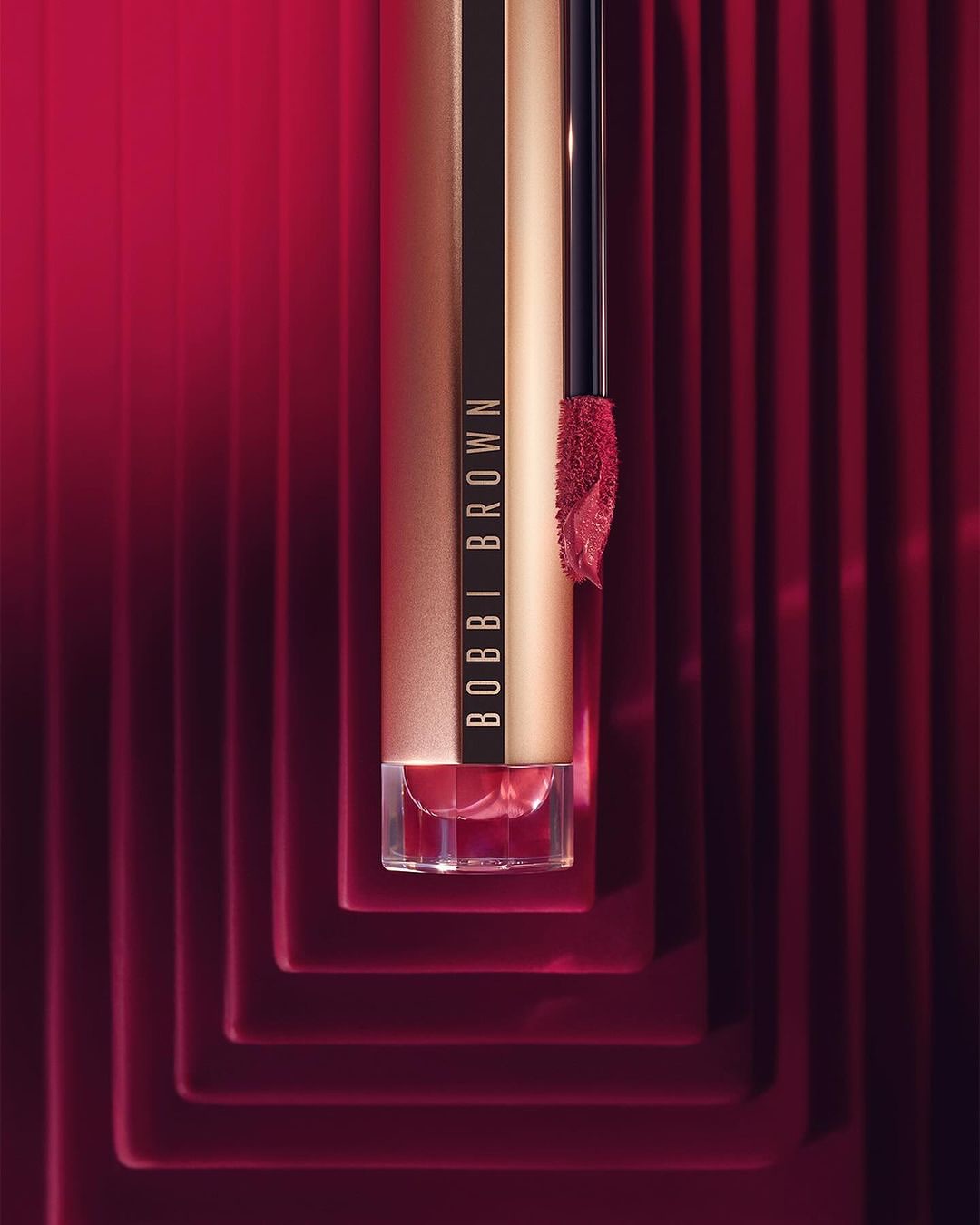 Close-up studio shot of Bobbi Brown lipstick in a luxurious gold tube, featuring the product’s applicator and bold red background.