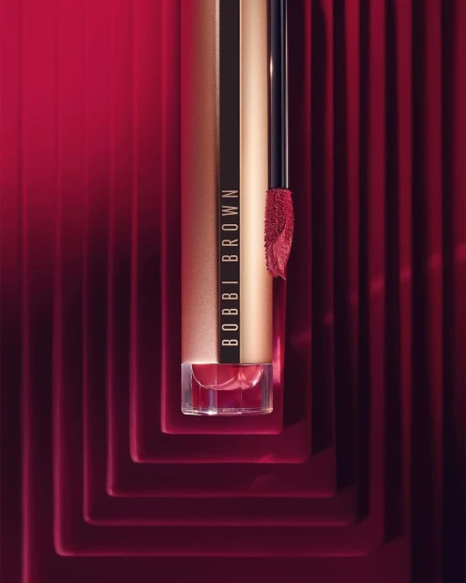 Close-up studio shot of Bobbi Brown lipstick in a luxurious gold tube, featuring the product’s applicator and bold red background.