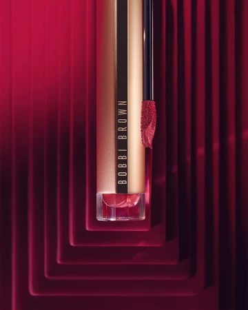 Close-up studio shot of Bobbi Brown lipstick in a luxurious gold tube, featuring the product’s applicator and bold red background.