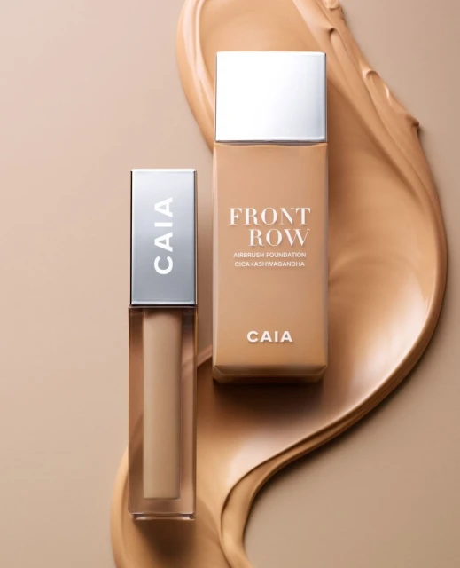 Two bottles of Caia Front Row airbrush foundation and concealer are displayed on a neutral background with a smear of foundation.