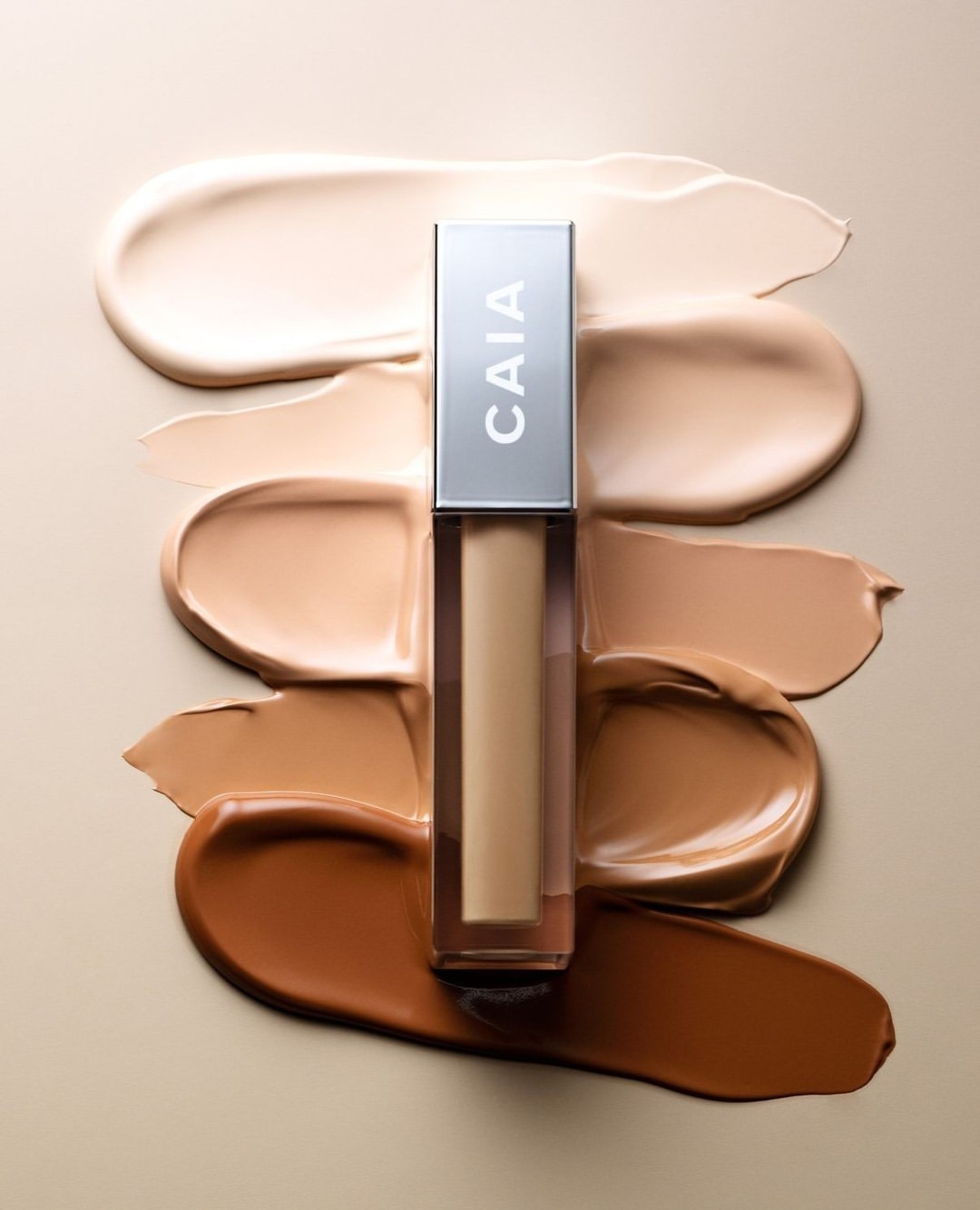 Cosmetic concealer tube by CAIA Cosmetics, positioned over color swatches of different skin tones on a beige background in a studio setting.