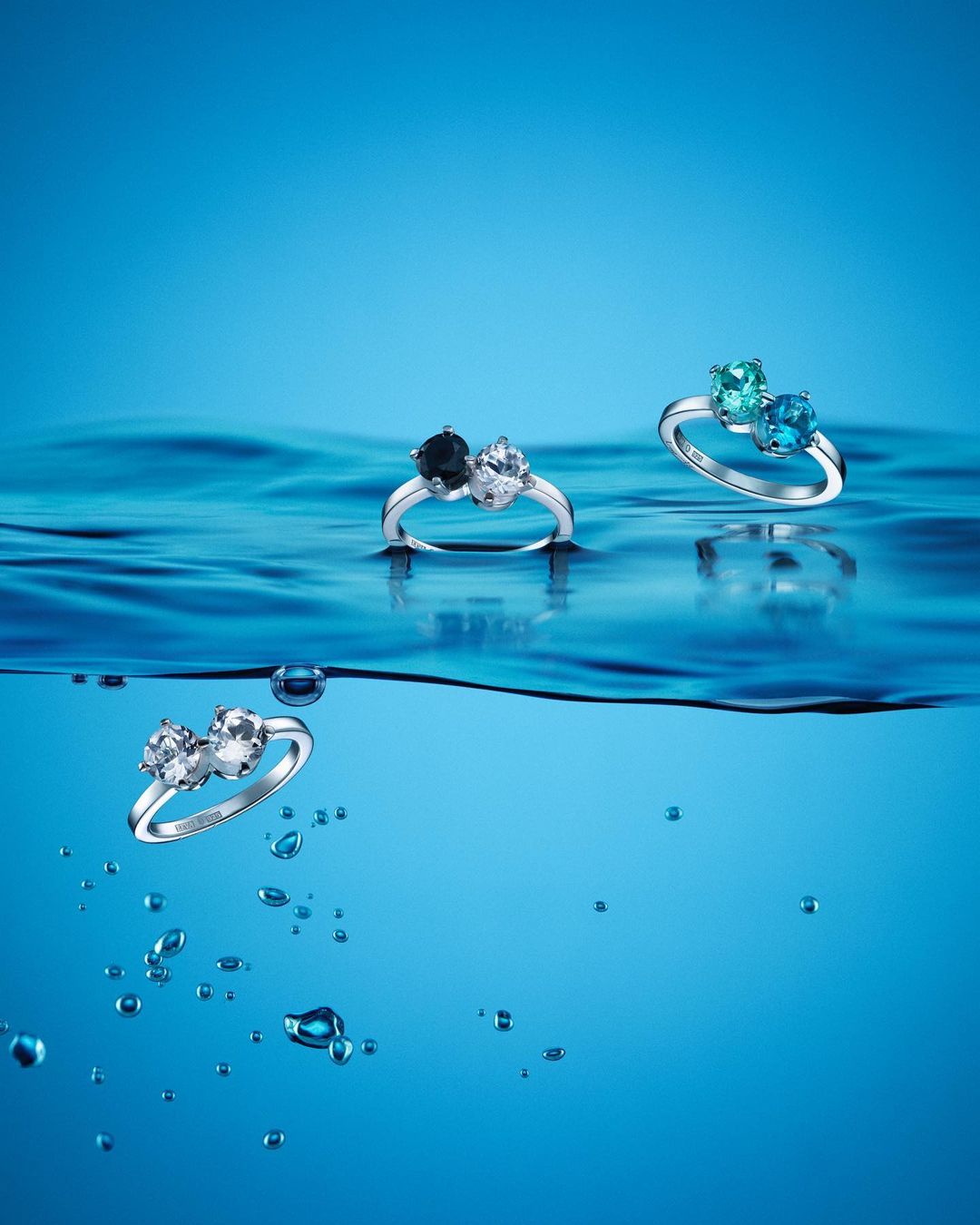 Three gemstone rings are displayed above the wavy surface of blue water in a product shot emphasizing elegance and color.