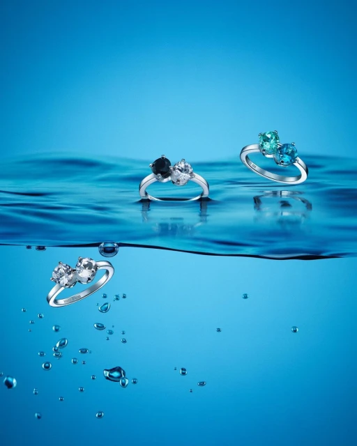 Three gemstone rings are displayed above the wavy surface of blue water in a product shot emphasizing elegance and color.
