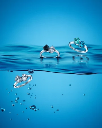 Three gemstone rings are displayed above the wavy surface of blue water in a product shot emphasizing elegance and color.