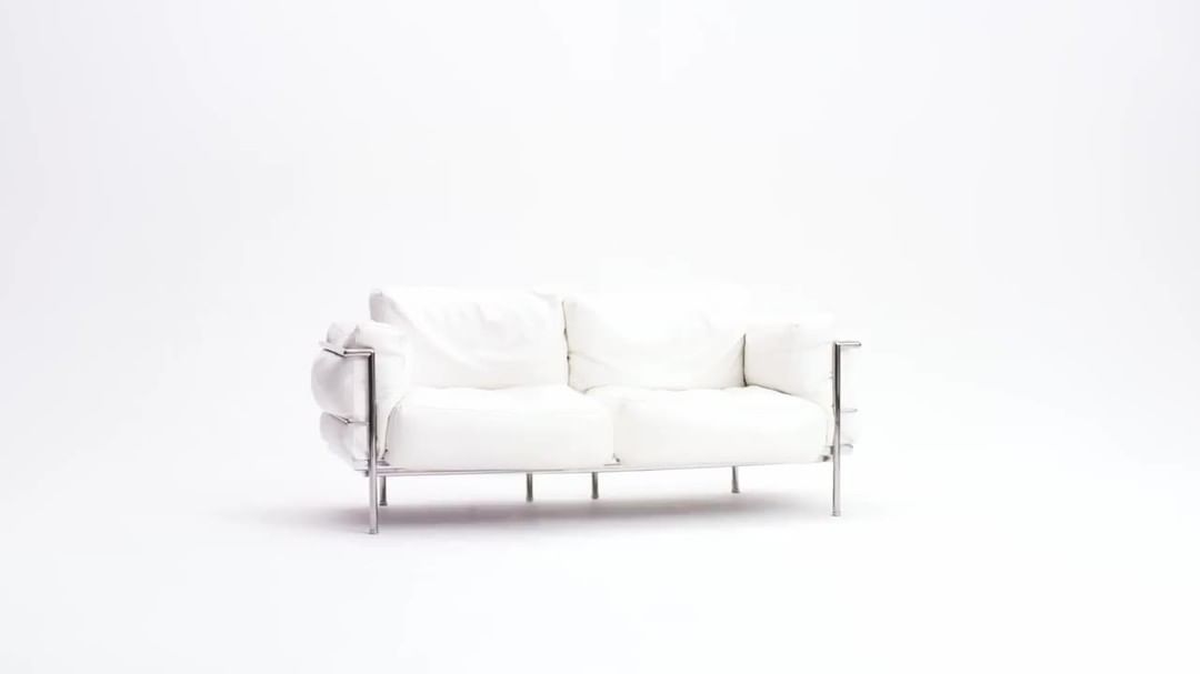 A two-seater white leather sofa with a chrome frame sits in the center of a minimalist white studio against a stark white background.
