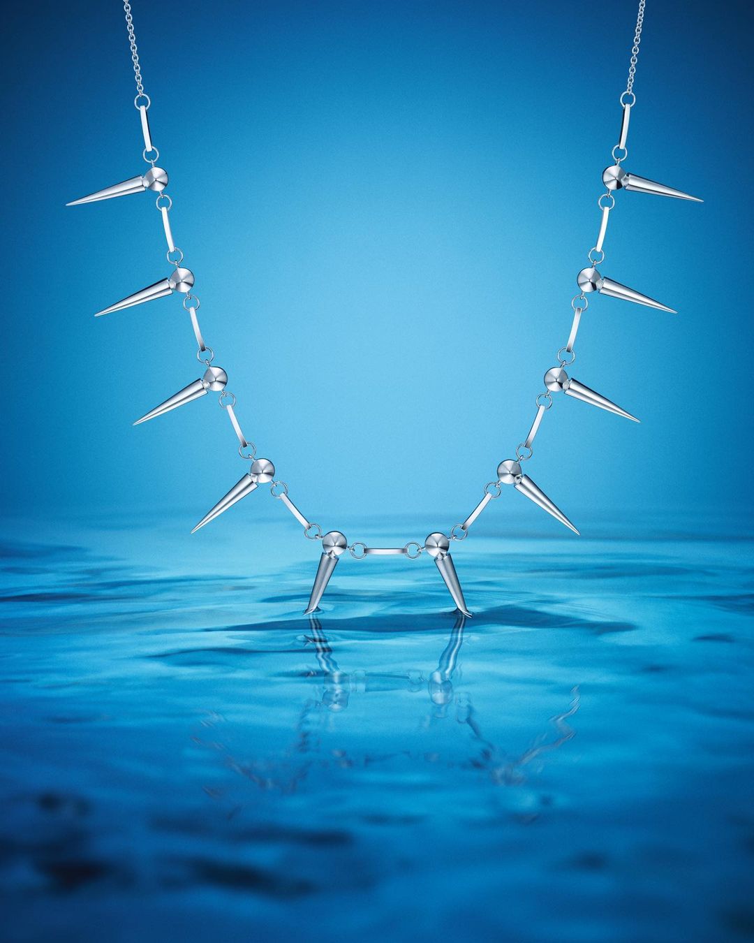 Silver spiked necklace suspended above rippling blue water against a matching blue background