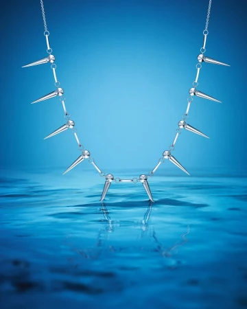Silver spiked necklace suspended above rippling blue water against a matching blue background