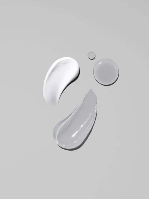 Swatches of facial cream in white and gray tones, along with two water droplets, showcased against a soft gray backdrop.