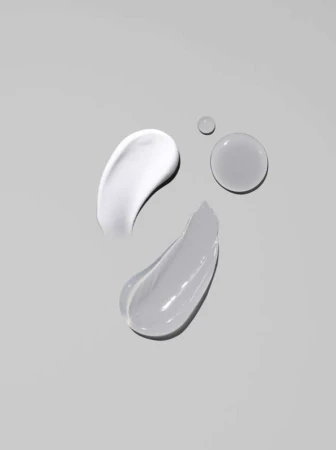 Swatches of facial cream in white and gray tones, along with two water droplets, showcased against a soft gray backdrop.
