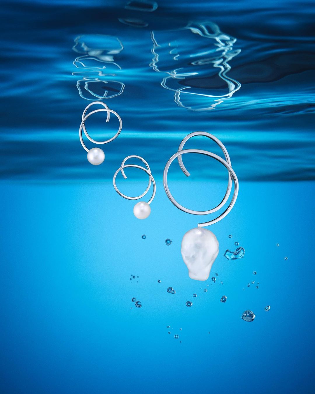 Elegant pearl and silver earrings under rippled water surface. Close-up jewelry shot with cool, blue water backdrop and floating water bubbles.