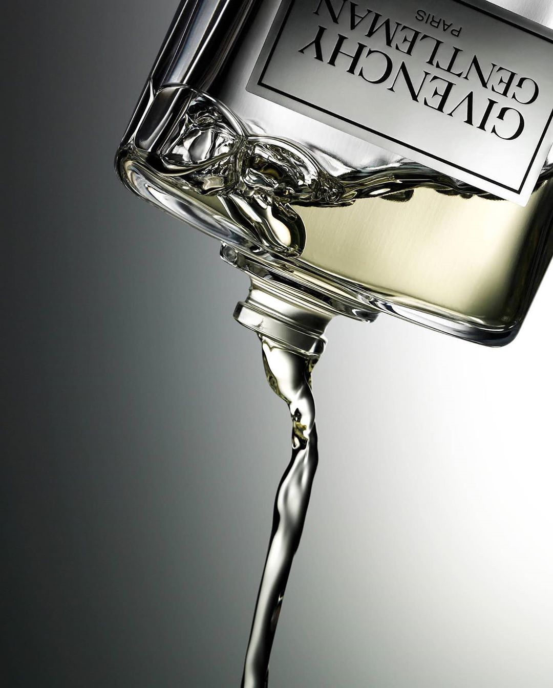 A bottle of Givenchy Gentleman fragrance tilted with liquid pouring out, set against a neutral gray gradient background in a beauty shot.