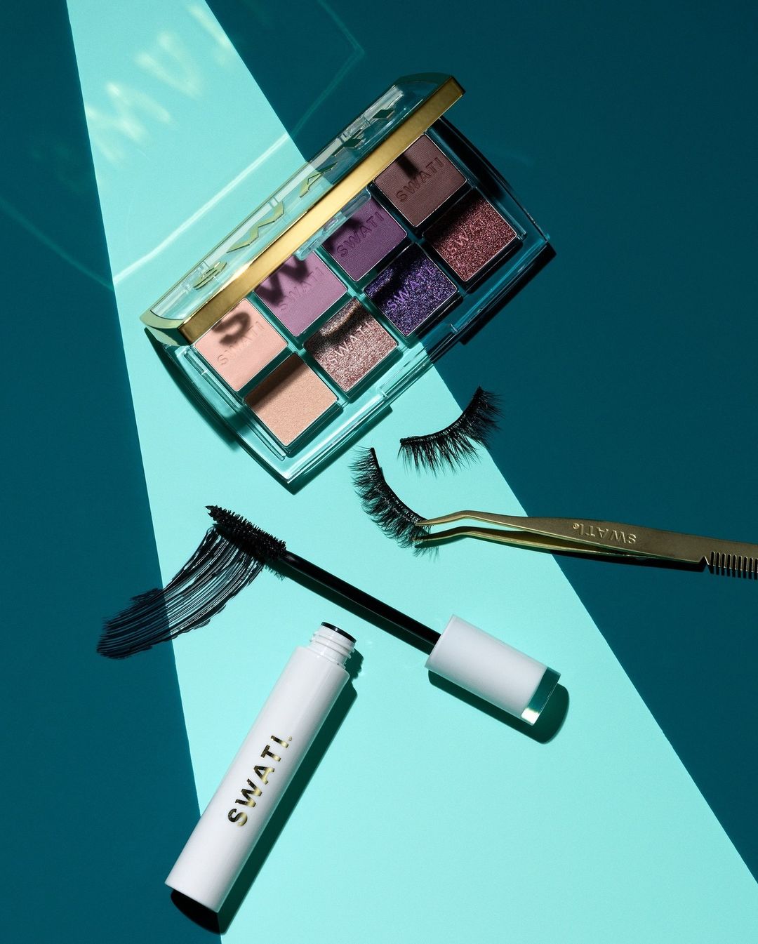 Cosmetics still life on a turquoise surface featuring an eyeshadow palette, mascara, false eyelashes, and gold tweezers.