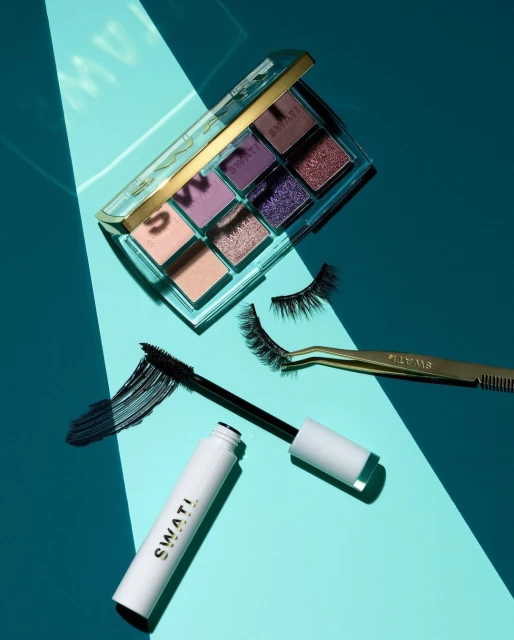 Cosmetics still life on a turquoise surface featuring an eyeshadow palette, mascara, false eyelashes, and gold tweezers.