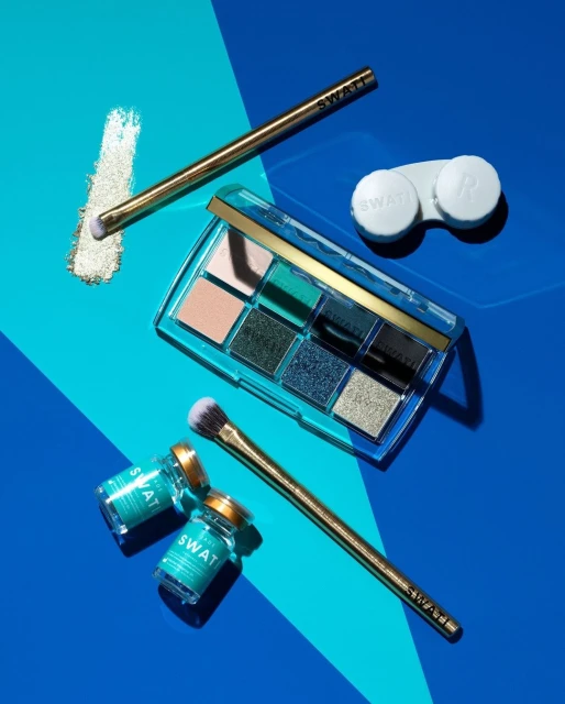 Eye shadow palette with various shades of color, two makeup brushes, two sample containers, and a contact lens case on a blue surface.