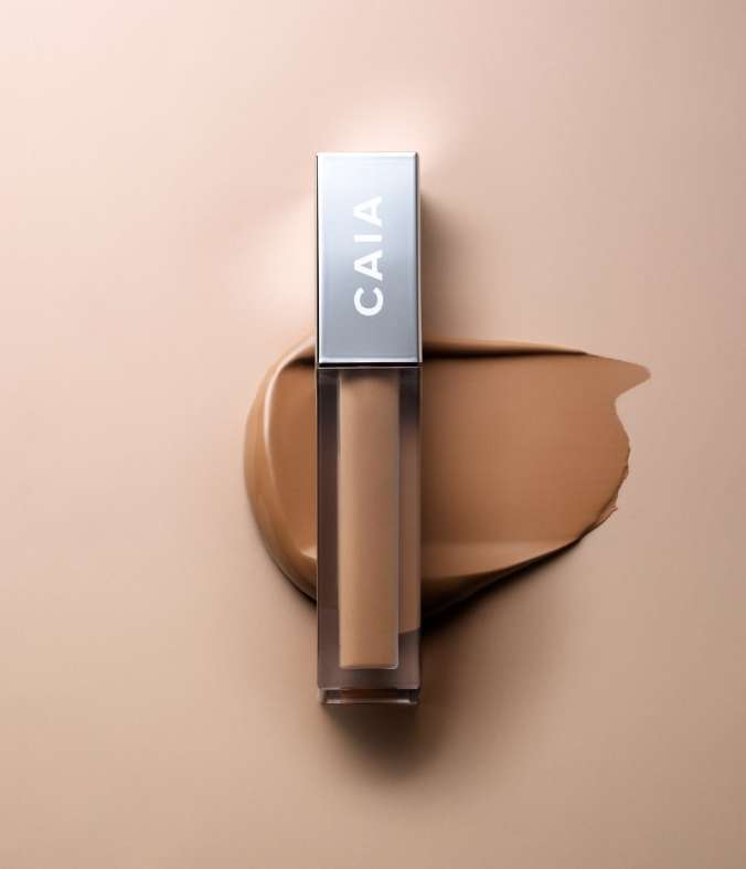 Close-up of a CAIA Cosmetics concealer tube lying on a beige surface next to a patch of concealer. Studio shot.