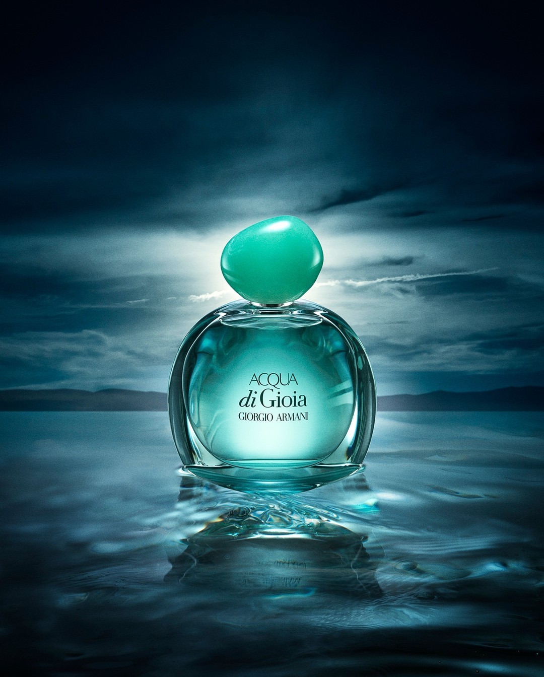 Aqua di Gioia fragrance bottle on reflective water surface with blurred cloudscape background in cool, blue-green tones. Beauty shot.