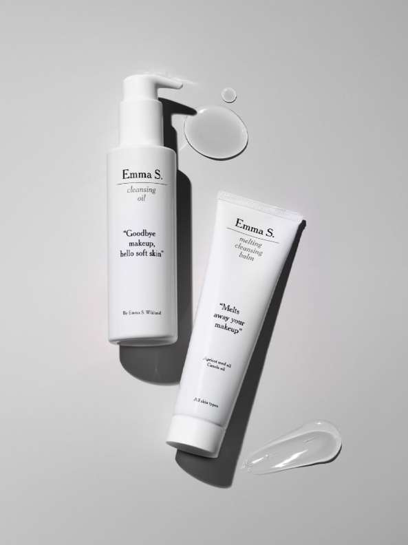 Two white bottles of Emma S. cleansing oil and melting cleansing balm on a gray surface with gel-like product smears nearby.