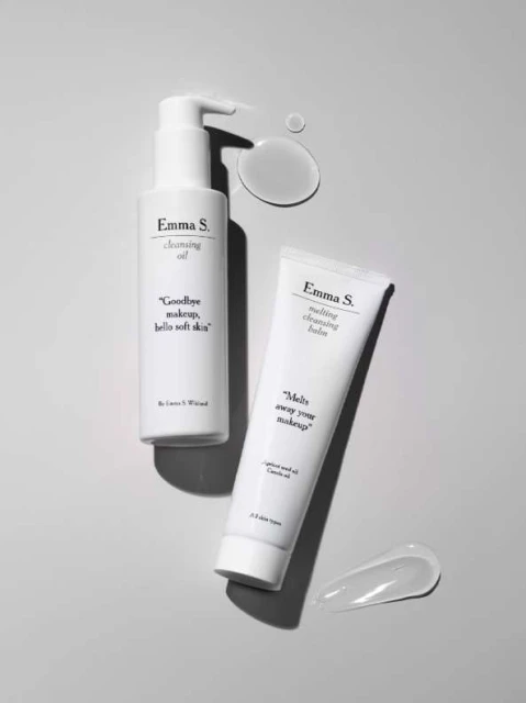 Two white bottles of Emma S. cleansing oil and melting cleansing balm on a gray surface with gel-like product smears nearby.