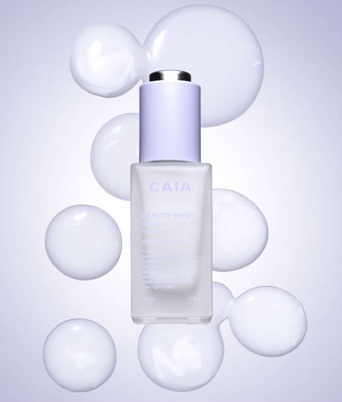 Cosmetic serum bottle with dropper top on a purple gradient background surrounded by white opaque spheres in a studio setting.