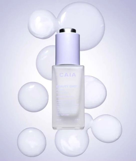 Cosmetic serum bottle with dropper top on a purple gradient background surrounded by white opaque spheres in a studio setting.