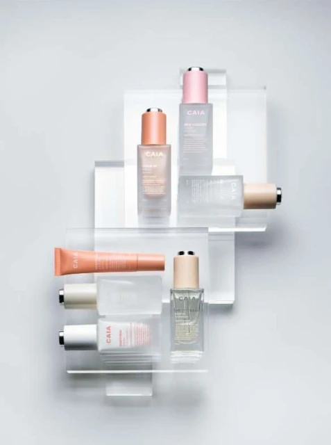 Elevated beauty product arrangement with CAIA cosmetics on clear acrylic platforms against a light gray backdrop in a studio setting.