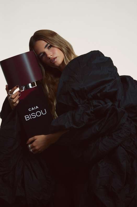 Brunette woman in ruffled black dress embraces a large perfume bottle, set against a pale background. The fragrance is 'CAIA BISOU'.